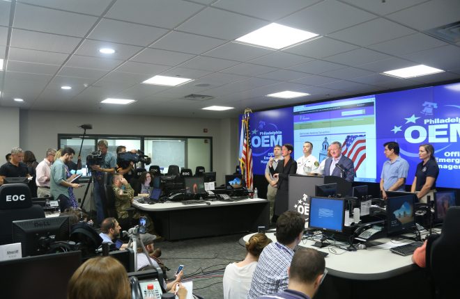 Reporters listen as Mayor Kenney speaks among computers and large screens displaying the OEM logo.