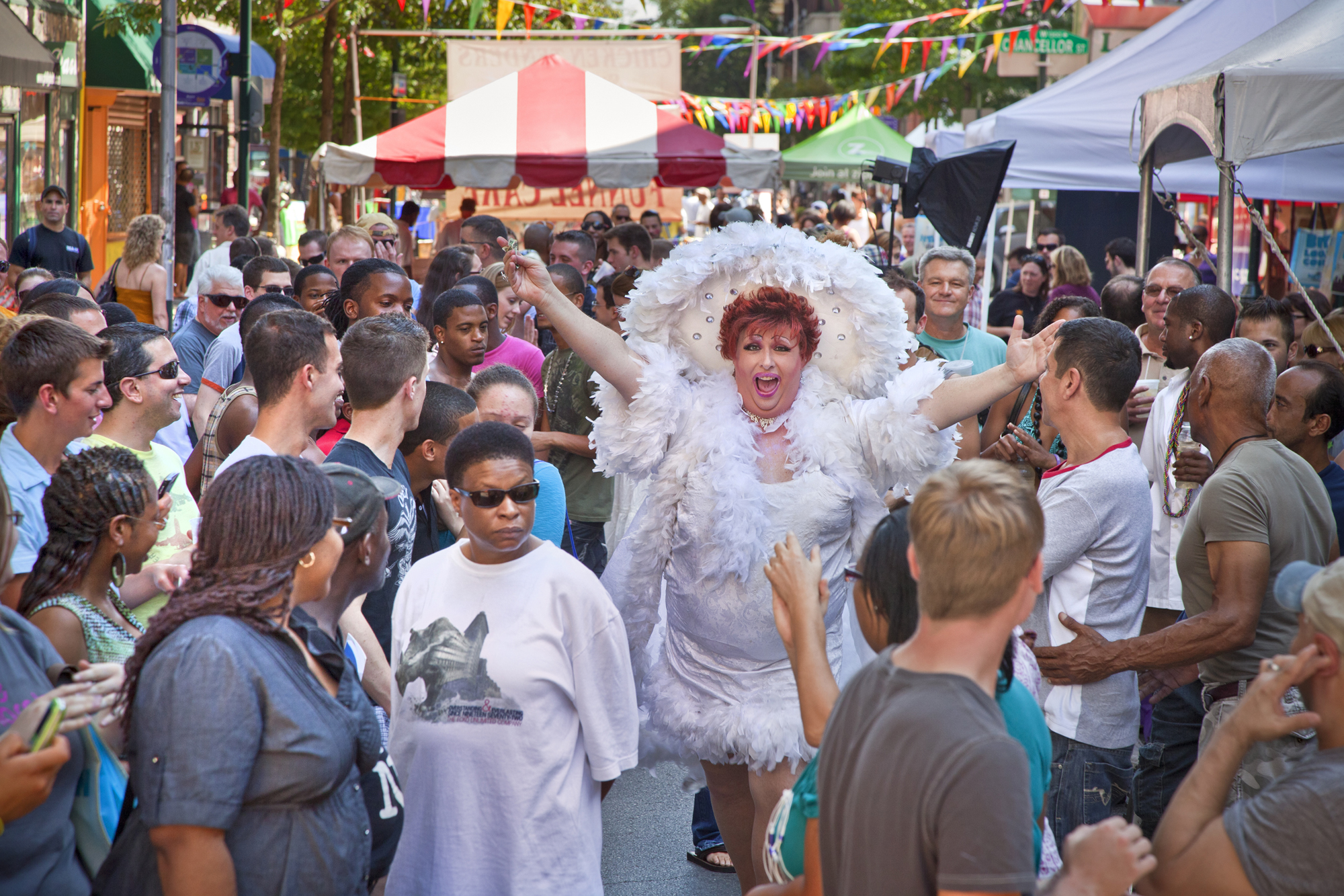 A closed off street filled with celebrants and vendors with a drag queen dressed in white with outstretched arms and a happy countenance.