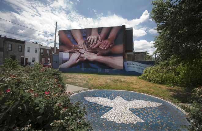 A mural depicts multicultural hands overlapping one another communicating peace, teamwork, and cooperation. The mural is on the side of a rowhouse in South Philadelphia. There is a community green space in front of it with bushes and a mosaic on a patio depicting a dove.