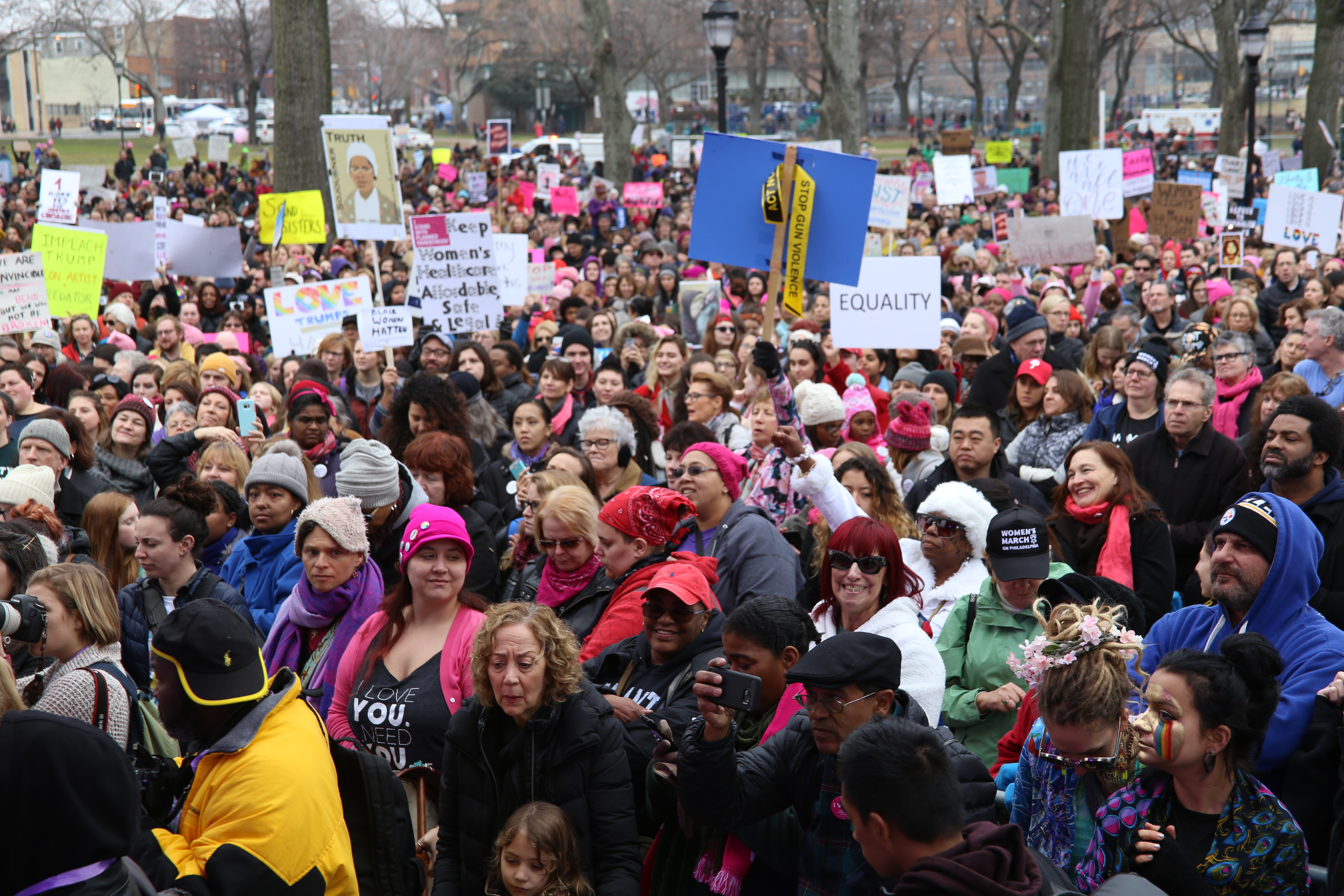 A crowd comprised mostly of women, some in pink scarves and hats, holding signs about equality and feminism. 