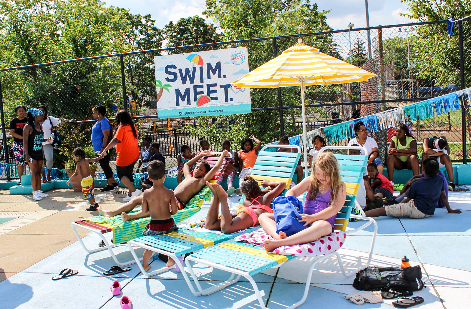 Child lounge on chairs near a pool and a Philadelphia Parks and Rec Swim Meet sign at a local rec center.