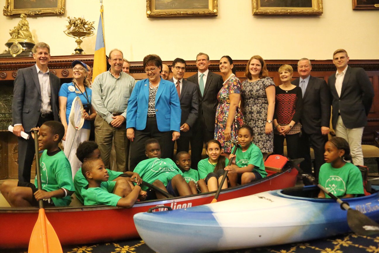 Children wearing matching t-shirts sit in a canoe in City Hall while the Mayor, City officials, and others stand behind them smiling.