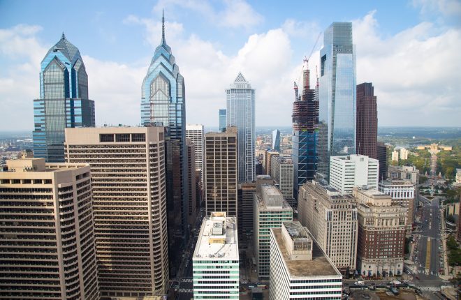 The City of Philadelphia's skyline as seen from City Hall's iconic clock tower. Glass skyscrapers reach toward a sky with several puffy clouds. It is a warm, sunny day, and the Philadelphia Museum of Art is far off in the distance.