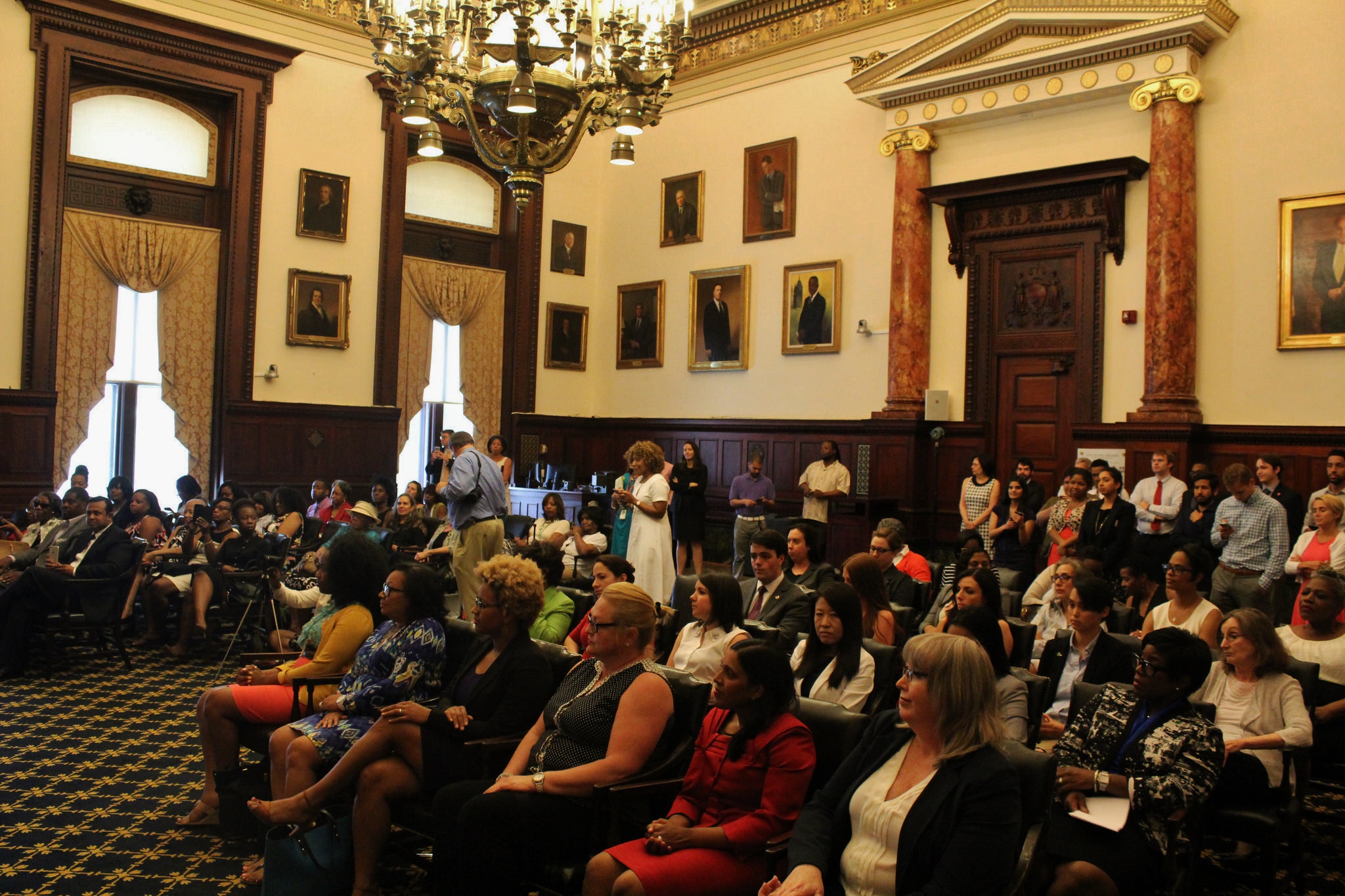 Women sit in the Mayor's Reception Room in City Hall. In the background there are paintings of former mayors.