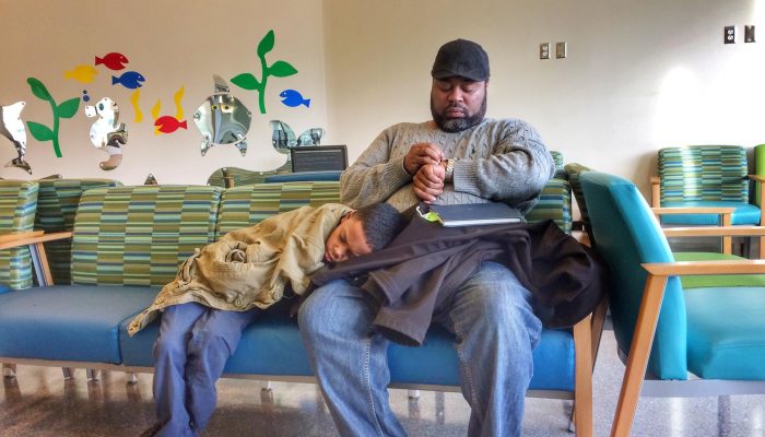 A father sits in a hospital waiting room with his young son resting on his lap.