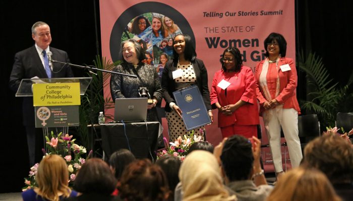 Mayor Jim Kenney speaks at a glass podium next to a group of women of all different backgrounds. There is a banner behind them that says the state of women and girls in Philadelphia. All are smiling and happy.