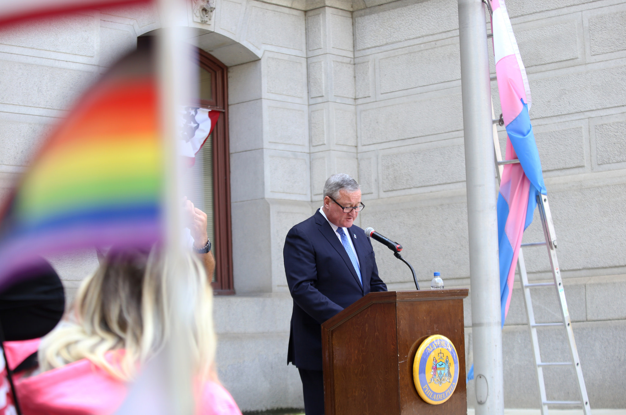 Mayor Kenney speaks in between an LGBTQ pride flag, with additional black and brown stripes representing people of color, and a trans pride flag at City Hall.