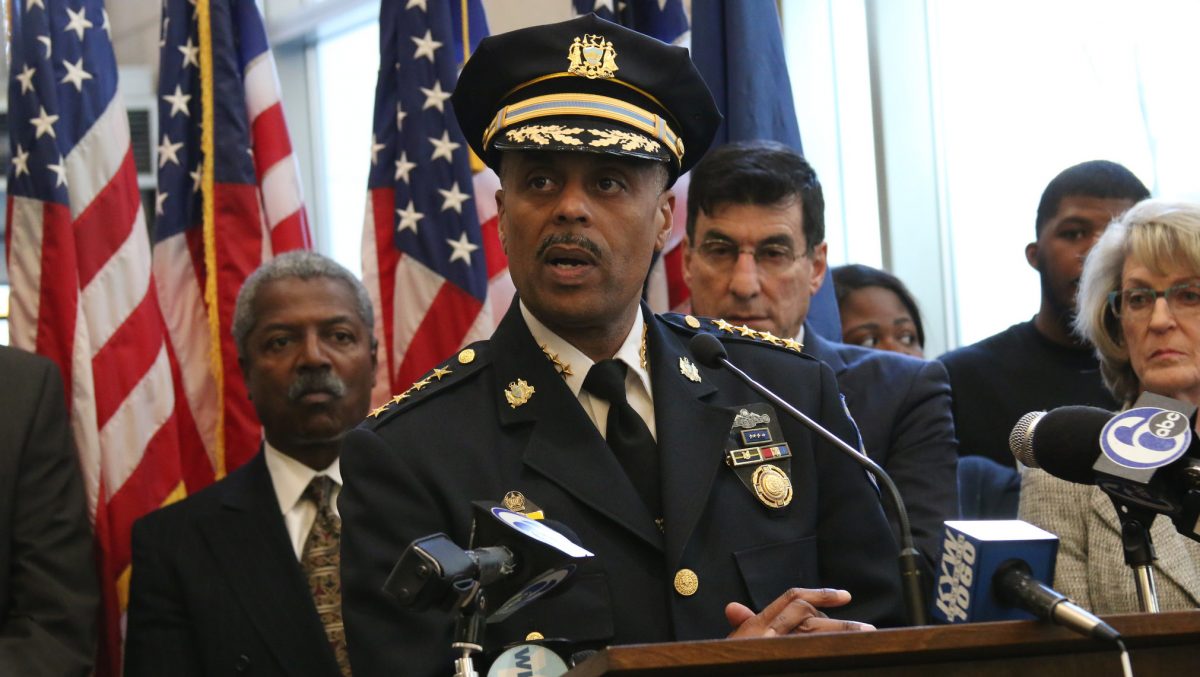 Wearing his uniform and hat, Commissioner Ross speaks at a podium. Behind him are several City officials and many American flags. Ross has a mustache.
