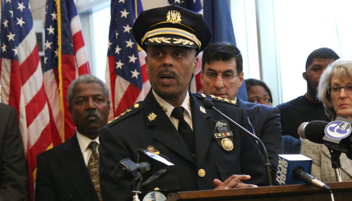 Wearing his uniform and hat, Commissioner Ross speaks at a podium. Behind him are several City officials and many American flags. Ross has a mustache.