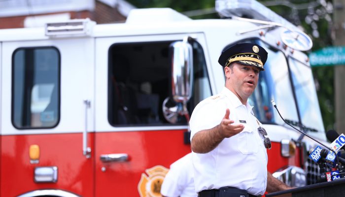 Commissioner Thiel stands at a podium speaking with a PFD engine behind him. He is wearing his officers cap and a short sleeve uniform shirt.