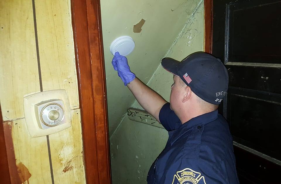 A firefighter reaches toward the ceiling, adjusting a round plastic smoke alarm attached to the ceiling above a staircase. He is wearing a Philadelphia Fire Department shirt and badge and PFD baseball cap.