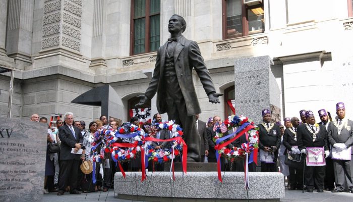 The Octavius V. Catto statue stands outside City Hall surrounded by patriotic themed wreaths. A diverse crowd of onlookers stands in the background. There are hundreds of people in and around the area.