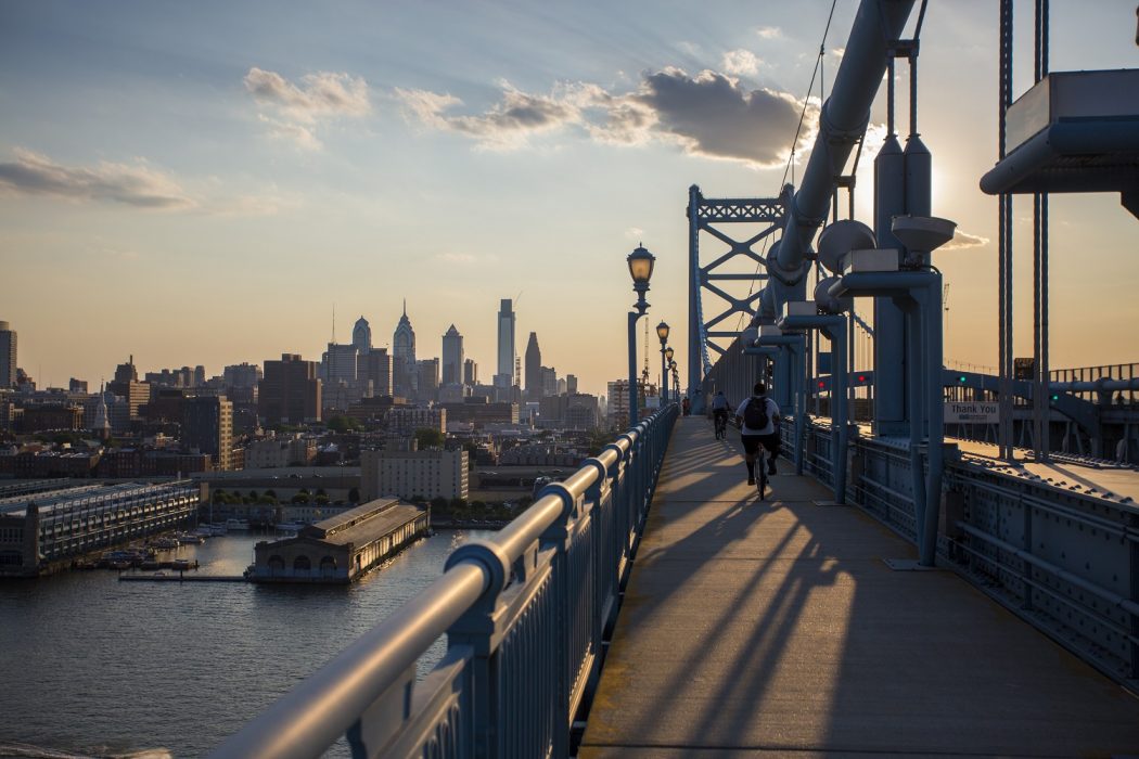 The skyline of Philadelphia rises on the horizon from the perspective of bicyclists and runners traversing the Ben Franklin Bridge. It is evening and the sky is clear with only a few puffy clouds. The image evokes possibility and beginnings.