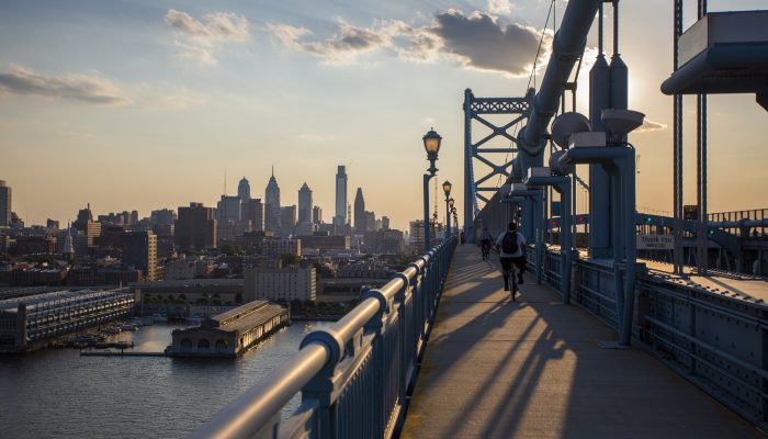 The skyline of Philadelphia rises on the horizon from the perspective of bicyclists and runners traversing the Ben Franklin Bridge. It is evening and the sky is clear with only a few puffy clouds. The image evokes possibility and beginnings.