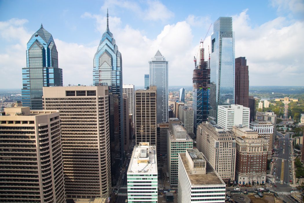 The City of Philadelphia's skyline as seen from City Hall's iconic clock tower. Glass skyscrapers reach toward a sky with several puffy clouds. It is a warm, sunny day, and the Philadelphia Museum of Art is far off in the distance.