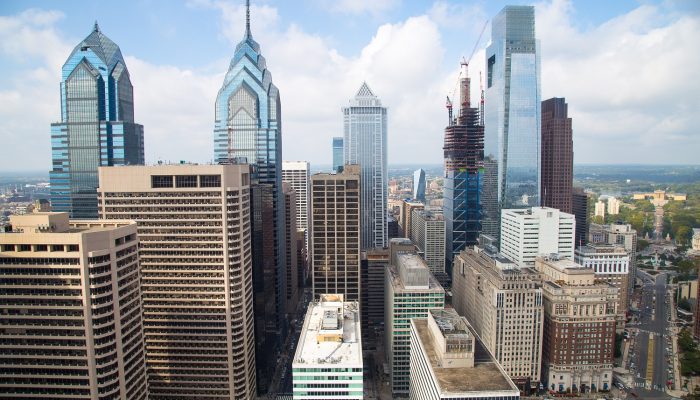 The City of Philadelphia's skyline as seen from City Hall's iconic clock tower. Glass skyscrapers reach toward a sky with several puffy clouds. It is a warm, sunny day, and the Philadelphia Museum of Art is far off in the distance.