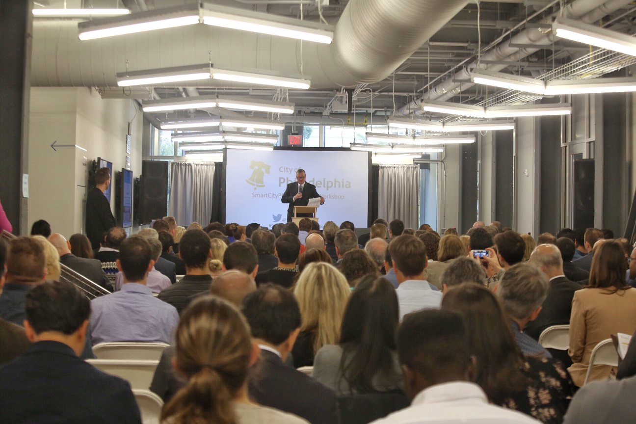 Mayor Kenney speaks at the far end of the room in front of a digital screen. There are dozens of people seated listening to him in a space that looks industrial with exposed duct work and steel lights hanging from the ceiling.