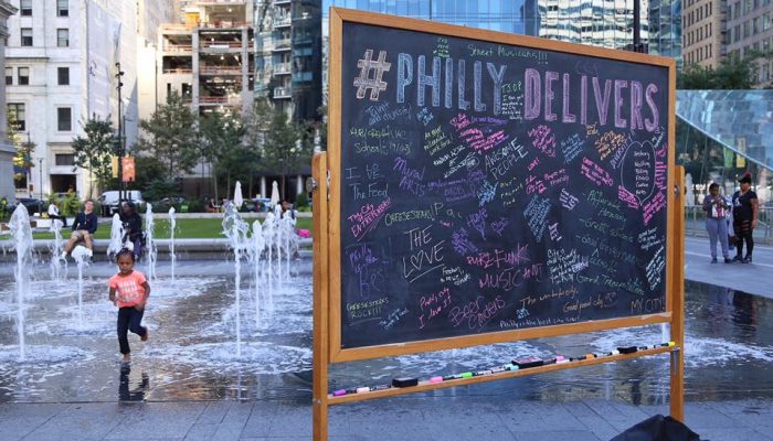 A chalkboard with dozens of different submissions all written by different people sits in Dilworth Park. Behind it, a child runs through the fountains playfully avoiding the water.