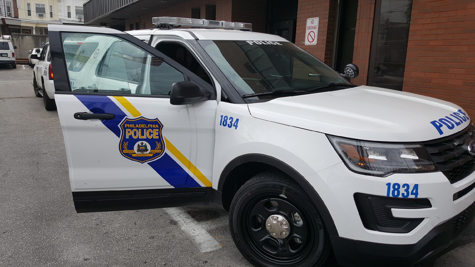 A sport utility style vehicle with the Philadelphia Police Department shield on it's open passenger front door sits in a police district parking lot. The vehicle is modern and has police lights on its roof.