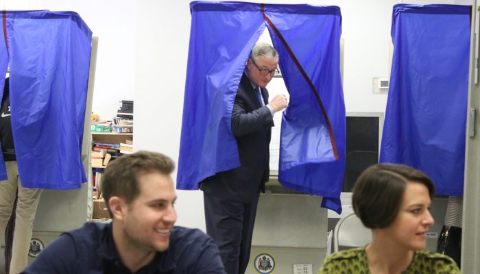 Mayor Kenney opens the curtain of a voting booth as he exits after he voted. There are two poll workers sitting in the foreground signing registered voters in.