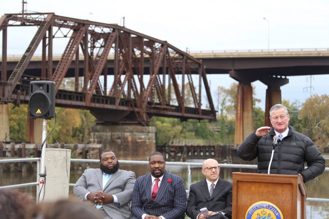 Mayor Kenny speaks at a podium in front of a huge industrial looking steel bridge. Councilman Kenyatta Johnson and State Representative Jordan Harris sit listening to him. alongside PIDC's John Grady. The Mayor is wearing a quilted, puffy winter coat.