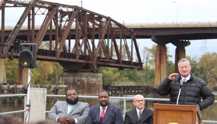Mayor Kenny speaks at a podium in front of a huge industrial looking steel bridge. Councilman Kenyatta Johnson and State Representative Jordan Harris sit listening to him. alongside PIDC's John Grady. The Mayor is wearing a quilted, puffy winter coat.