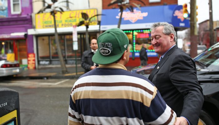Mayor Kenney smiles as he shakes hands and grabs the elbow of a neighbor. Behind them are inviting small businesses with electronic signs.
