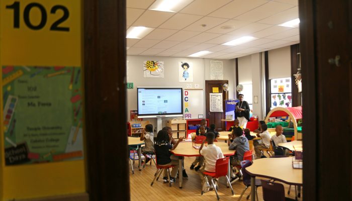 Young students sit in little chairs at kid-sized tables listening to a teacher. The classroom's door and threshold frames the picture as though you are standing in the doorway. There are kids' projects on the walls and big letters as though they're learning the alphabet.