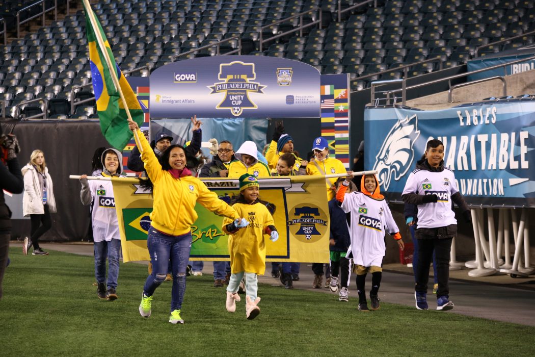 Brazilians of all ages, wearing colors that mimic the colors of the Brazilian flag, proudly walk their banner out onto the field. Smiling, they excitedly wave the Brazilian flag.
