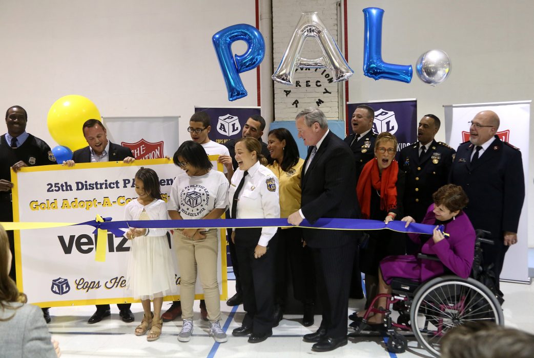 A line of diverse people and kids stands as a child cuts a ribbon with a large pair of scissors. There are traditional round balloons and lettered balloons spelling PAL.