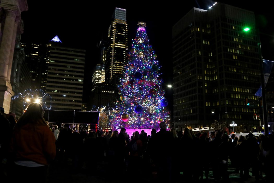 A large diverse crowd gathers at night. In awe, they admire the lit Christmas tree in Dilworth Park. Behind the tree stands skyscrapers and other buildings of the Philly skyline.