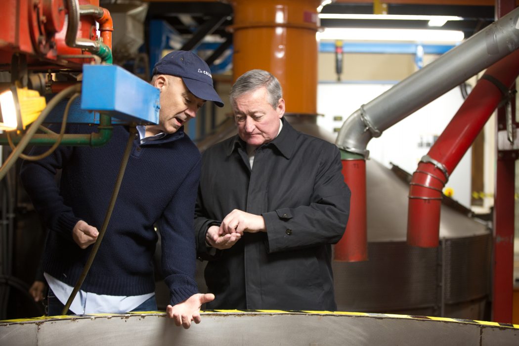 Mayor Kenney holds a handful of coffee beans as he looks into a large metal machine. There are big industrial pipes in the background, and Todd Carmichael is wearing a La Colombe hat as he points at the machine they're both standing in front of.