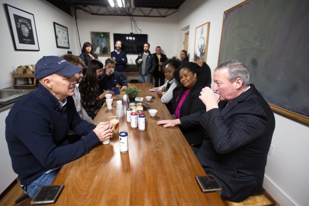 Mayor Kenney sits at a long table with staff from his office and La Colombe. He's sipping coffee as Todd Carmichael speaks energetically. Carmichael is wearing a sweater and a La Colombe cap; the Mayor is wearing a suit and a winter coat.