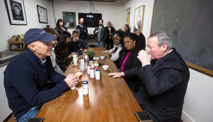 Mayor Kenney sits at a long table with staff from his office and La Colombe. He's sipping coffee as Todd Carmichael speaks energetically. Carmichael is wearing a sweater and a La Colombe cap; the Mayor is wearing a suit and a winter coat.