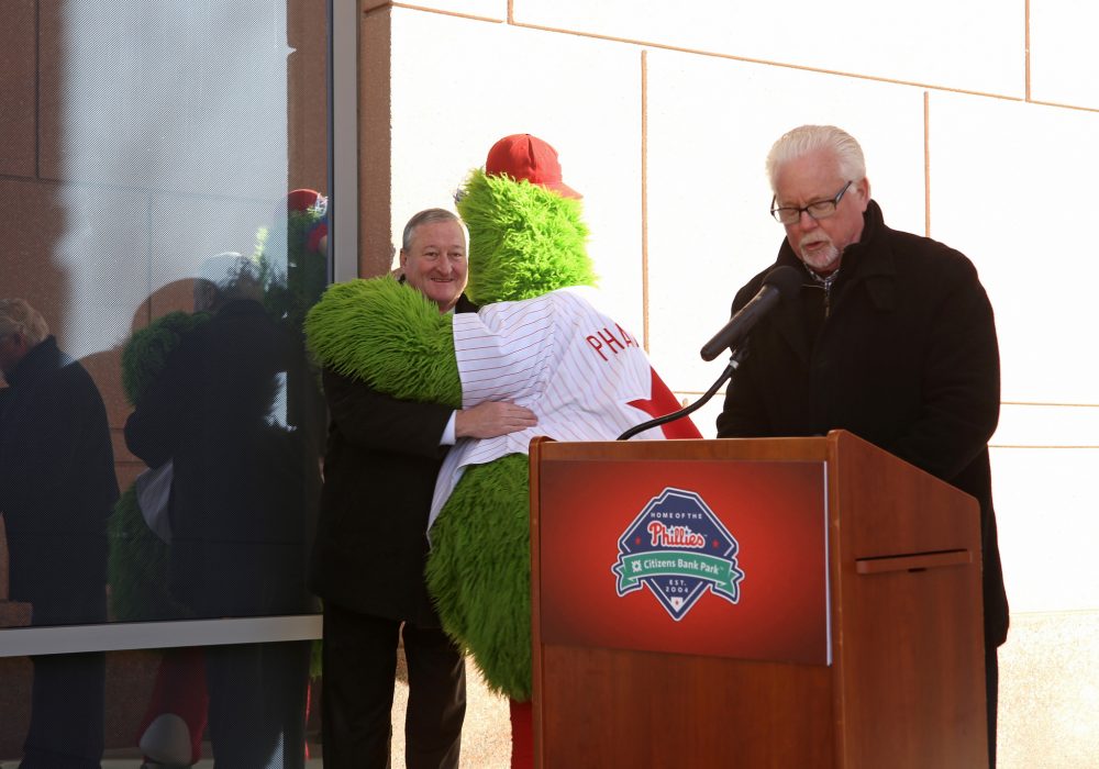 A man speaks at a podium as the Phillie Phanatic tightly hugs Mayor Kenney. The Mayor looks very happy like he's hugging an old friend.