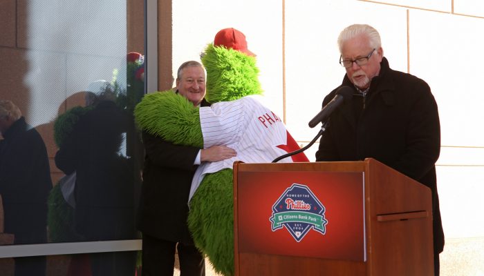 A man speaks at a podium as the Phillie Phanatic tightly hugs Mayor Kenney. The Mayor looks very happy like he's hugging an old friend.