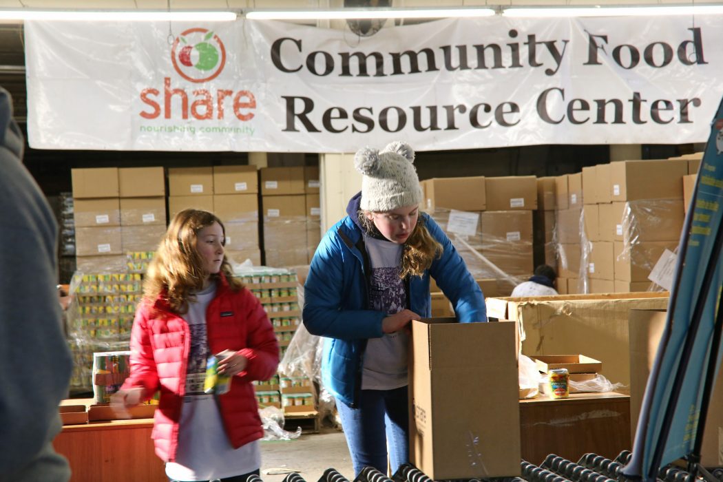 Two girls pack paper bags of food. Behind them are stacks and stacks of boxes of dry and canned goods. There's a banner that says, "Community Food Resource Center" in the background. The girls are dressed in winter coats and one has a wool cap on.