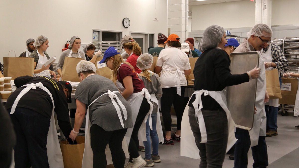 Volunteers wearing aprons and hair nets move cardboard boxes and paper bags around an industrial kitchen at MANNA. They are preparing meals to be delivered.
