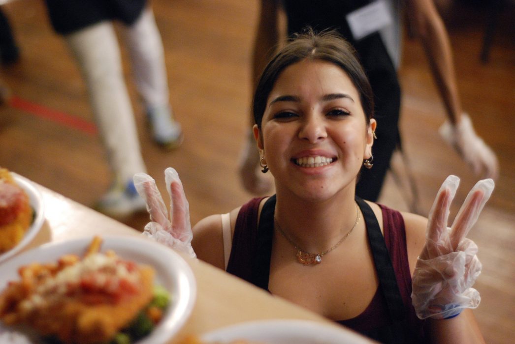 A woman wearing plastic gloves gives the peace sign on both hands and smiles. She's standing in front of a table of plates of food to be distributed at free meal site here in Philadelphia.