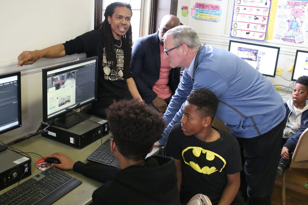 Mayor Jim Kenney wears his glasses as he peers at a computer screen. On the screen is the Coded by Kids website. An instructor stands with his arm on the monitor and two high school aged boys are seated at the computer.