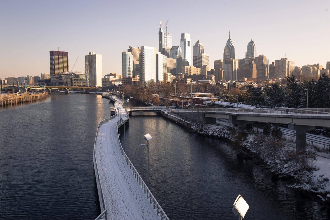 Iconic skyscrapers that make up Philly's skyline are on the horizon about a mile away. The perspective is from standing on the South Street Bridge. There is a boardwalk in the Schuylkill River covered in snow. The sky is clear and it is morning.