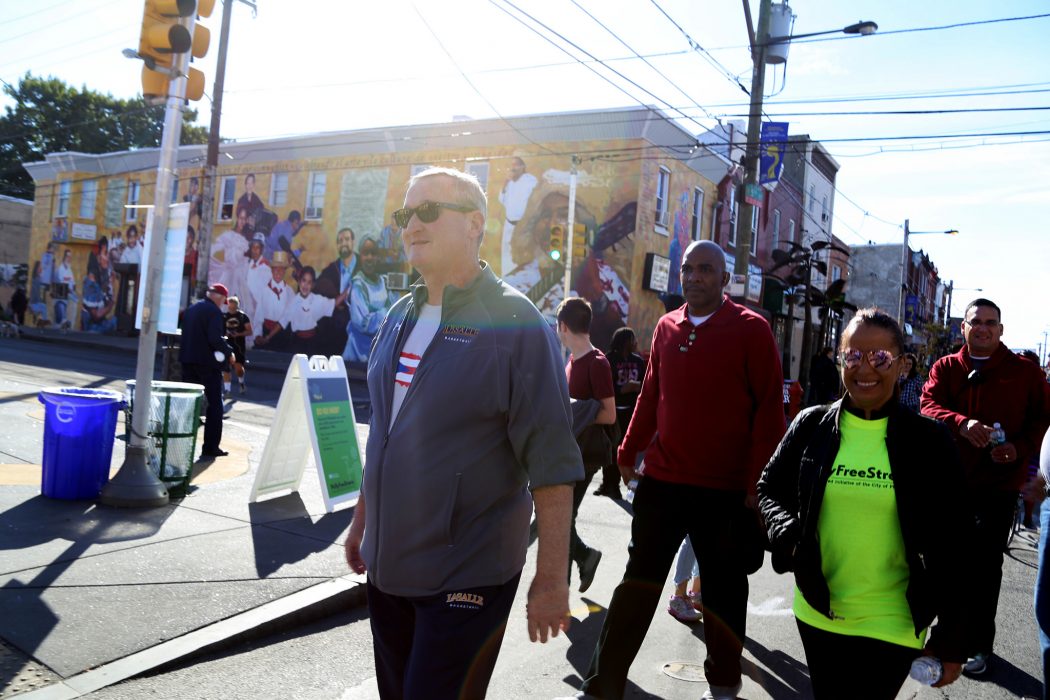 Mayor Kenney walks in a sweat suit and a t-shirt with a dozen happy, smiling people walking behind him. They are walking in the middle of the street because the streets are closed down so people can walk, bike, dance, and stroll for Philly Free Streets. The sun is out and it is warm even though it's autumn.