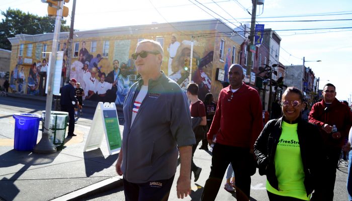 Mayor Kenney walks in a sweat suit and a t-shirt with a dozen happy, smiling people walking behind him. They are walking in the middle of the street because the streets are closed down so people can walk, bike, dance, and stroll for Philly Free Streets. The sun is out and it is warm even though it's autumn.