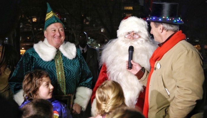 Mayor Kenney wears a costume like Will Farrell's character in the movie "Elf." It is made of velvet and he is wearing a point hat with a big leather belt and gold belt bucket. He has fur cuffs and a collar. He is standing next to Santa Claus who has a big bushy beard and glasses on. There are happy children nearby, and a journalist is interviewing Santa Claus.