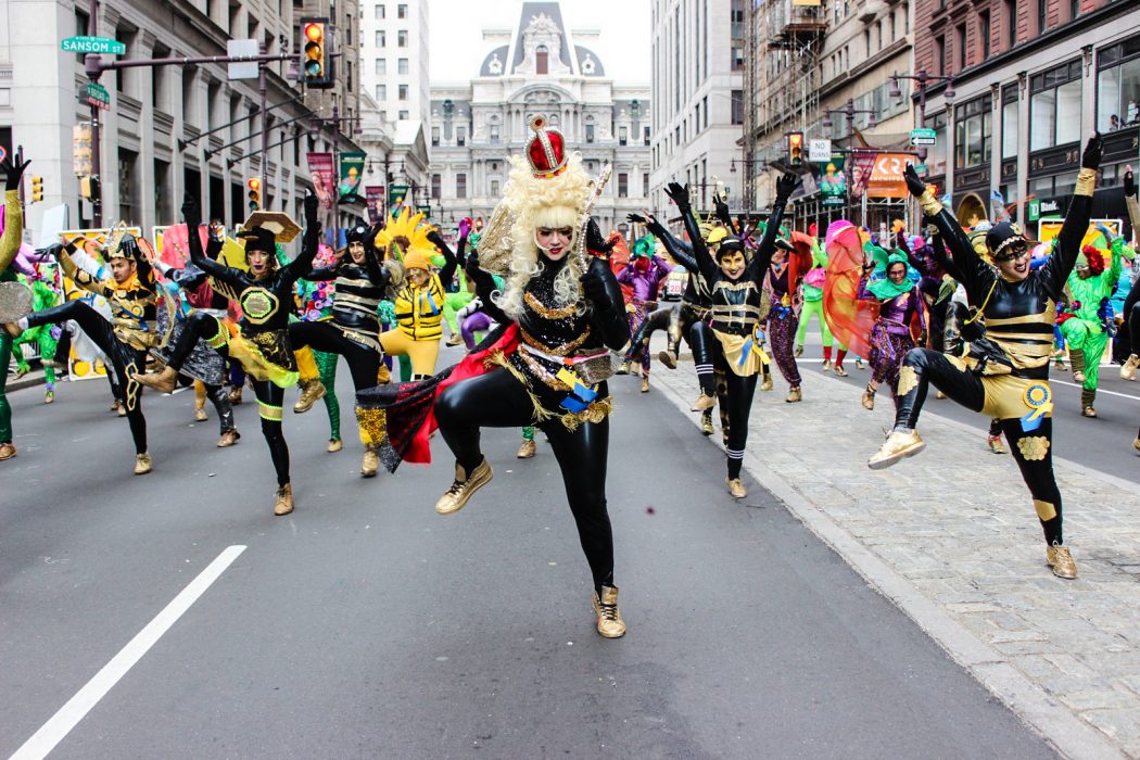Dozens of people dance on Broad Street in front of City Hall during 2016's Mummer's Parade. They are wearing big wigs and sequined, bejeweled costumes. They look happy and are dancing in a coordinated way with their legs and arms shooting into the air.