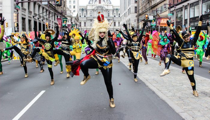 Dozens of people dance on Broad Street in front of City Hall during 2016's Mummer's Parade. They are wearing big wigs and sequined, bejeweled costumes. They look happy and are dancing in a coordinated way with their legs and arms shooting into the air.
