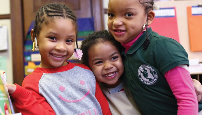 Three girls hug put their arms around one another and smile at the camera.