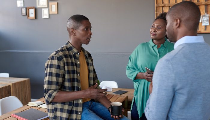 A man sits on a table holding a cup of coffee and talks to two other people.