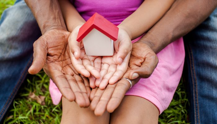 A large set of black hands and a small set of white hands hold a small wooden house.