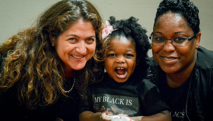 A little girl sits between two women. All three smile at the camera.